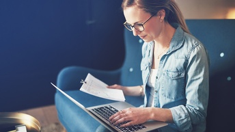 Person mit Brillen sitzt auf blauem Sofa und hält in einer Hand Dokumente, mit der anderen Hand tippt sie auf Laptoptastatur