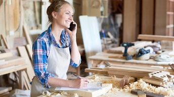 Person mit kariertem Hemd und Arbeitsschürze telefoniert freudig und macht sich Notizen auf einem Klemmbrett während sie in einer Tischlerei steht