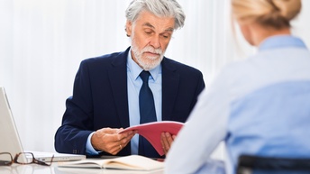 Eine Person schaut die Bewerbungsmappe einer zum Bewerbungsgespräch eingeladenen Person durch, die gegenüber von ihr sitzt.