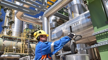 Person in blauem Overall mit gelbem Helm und Schutzbrillen dreht an Rad einer Gasanlage, ringsum verschiedene Leitungen und Container