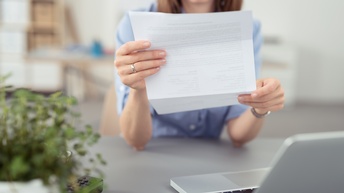 Nahaufnahme einer Person mit braunen Haaren, die konzentriert auf ein Dokument aus Papier blickt, das sie in beiden Händen hält. Sie steht und hat die Arme auf einen grauen Tisch gestützt, auf dem ein aufgeklappter Laptop und eine Topfpflanze steht