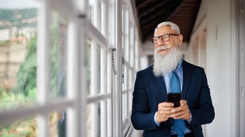 Lächelnde Person mit langem weißen Bart und Brillen hält Smartphone in Händen und blickt aus Fenster