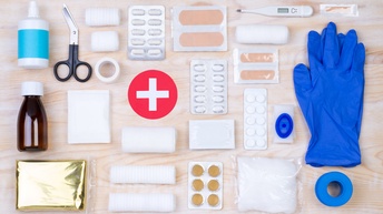 Auf einem hellen Holzuntergrund sind Objekte zum Thema Erste Hilfe wie Medikamente, Schutzhandschuhe, Scheren, Verbandsmaterial, Fiebermesser sowie eine Schere und diverse Fläschchen gelistet, Topshot
