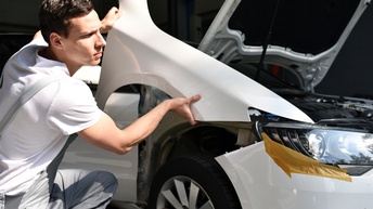 Eine Person in grauer Latzhose ist knapp neben einem weißen Auto, dessen Motorhaube geöffnet ist. Die Person hält ein weißes Teil auf das Auto. Beim vorderen Scheinwerfer des Autos ist ein braunes Klebeband