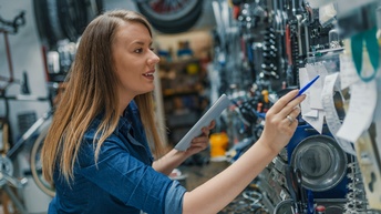 Person mit langen braunen Haaren steht in einer Werkstätte, hält ein Tablet in der Hand und blickt dabei auf aufgehängte Notizen