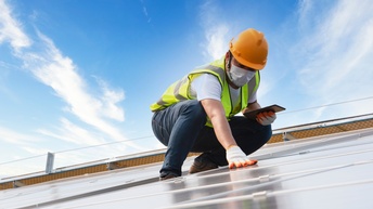 Person mit orangem Schutzhelm, gelber Warnweste, Mund-Nasen-Schutz und Tablet steht auf einem Dach und kontrolliert Solarpanele mit Schutzhandschuhen, im Hintergrund zeigt sich ein blauer Himmel mit Wolken