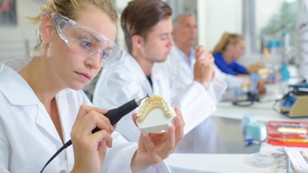 Person mit Schutzbrille und weißer Arbeitskleidung arbeitet an einer Zahnprothese mit einem elektrischen Schleifgerät, im Hintergrund sitzen weitere Personen an Arbeitsplätzen