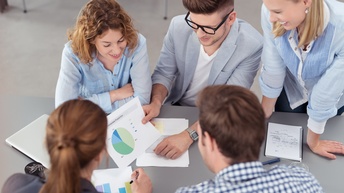 Eine Gruppe aus Personen sitzt und steht rund um einen Tisch und blickt auf verschiedene Unterlagen, Vogelperspektive