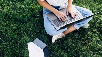 Eine Person in Jeans und Turnschuhen sitzt im Schneidersitz auf einer grünen Wiese. Die Person hat am Schoß einen aufgeklappten Laptop, auf dem sie tippt. Neben ihr liegen zwei Notizblöcke und ein Handy