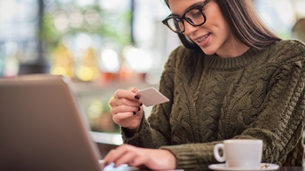 Person mit Brille und grünem Strickpullover sitzt mit einer Tasse vor einem Laptop und hält eine EC-Karte in der Hand während im Laptop etwas eingegeben wird