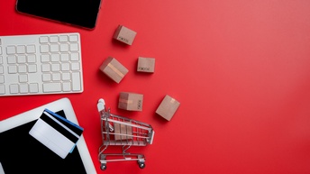 Auf einem roten Untergrund liegen in der linken Hälfte ein Smartphone, eine weiße Tastatur, ein Tablet, auf dem zwei Karten liegen, kleine Kartons und ein kleiner Einkaufswagen