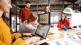 Mehrere Personen in Besprechungsraum um Tisch stehend und sitzend vor Computermonitoren, auf denen Analysen und Statistiken zu sehen sind
