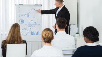 Person malt mit Stift auf Flipchart Diagramme, im Vordergrund mehrere Personen sitzend in Rückenansicht