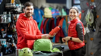 Zwei Personen stehen sich an einem Tresen in einem Geschäft gegenüber. Auf dem Tresen liegen ein Rucksack und ein Kletterhelm. Die linke Person hält eine Flasche. Die Personen lächeln und blicken in die Kamera