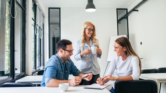 Personen mit Brillen amüsieren sich freudig mit einer Tasse sowie einem Notizbuch mit Stiften und sitzen und stehen in einem Raum mit mehreren weißen Tischen und schwarzen Sesseln, links befinden sich große Fenster und der Raum ist hell und freundlich