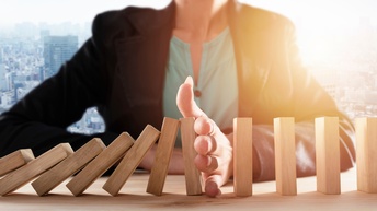 Die Büste einer Person ist zu sehen, sie sitzt an einem Holztisch. Mit der Hand stößt sie rechteckige Bausteine um. Im Hintergrund sieht man Umrisse einer Großstadt mit Hochhäusern. 