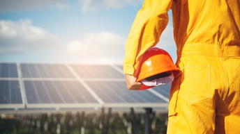 Rückenansicht Detail einer Person in gelbem Arbeitsoverall mit orangem Helm in Hand, im Hintergrund verschwommen Solarpanele