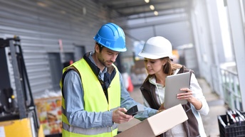 Frontalaufnahme zweier nebeneinander stehender Personen mit Helmen. Die linke Person hält ein Paket. Die rechte Person hält ein Tablet. Im Hintergrund sind die Außenwand einer Lagerhalle und ein Gabelstapler