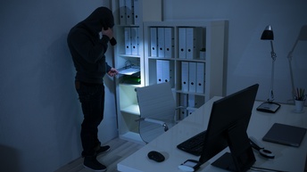 Eine schwarz gekleidete Person mit Sturmhaube ist in einem schlecht ausgeleuchteten Raum mit einem Schreibtisch. Sie hält in der rechten Hand eine Taschenlampe, mit der sie einen Aktenordner beleuchtet, der in einem Regal mit anderen Ordner steht