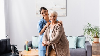 Zwei lächelnde Personen, die eine in blauem Arbeitsgewand legt Hände auf Schultern der anderen, die sich auf Stock abstützt