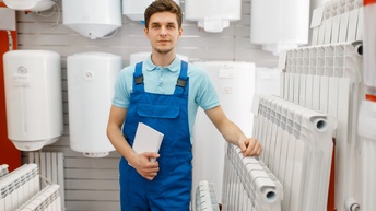 Person mit kurzen Haaren, Bart, blauem Polo-Shirt und blauer Latzhose steht in einem Ausstellungsraum mit Wasserboilern und Heizkörpern, lehnt  sich an und hält einen Notizblock in Händen