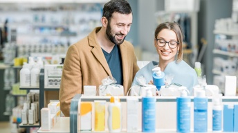Zwei Personen stehen in einem Geschäft nebeneinander an einem niedrigen Regal. Auf dem Regal stehen verschiedene Kosmetikartikel. Die rechte Person hält eine Flasche. Beide Personen blicken darauf