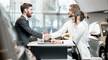 Drei lächelnde Personen in Autohaus stehend, zwei Personen reichen sich über Tisch die Hand