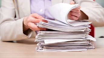 Vor einer Person steht auf einem Tisch ein Papierstapel. Die Person blättert durch den Stapel