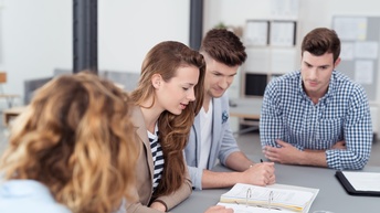 An einem Tisch sitzen vier Person. Auf dem Tisch liegen Unterlagen sowie ein aufgeklappter, dicker Ordner mit vielen Zetteln. Die Personen blicken auf die Zettel im Ordner