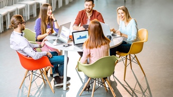 Fünf Personen sitzen auf bunten Sesseln um einen runden Tisch herum. Sie haben Laptops aufgeklappt, auf denen Diagramme zu sehen sind. und lächeln sich gegenseitig an.