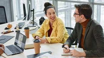Zwei Personen an Arbeitstisch sitzend in Unterhaltung miteinander, am Tisch verschiedene Arbeitsutensilien, Dokumente mit statistischen Grafiken und Kaffeebecher