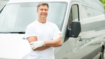 Lächelnde Person in weißer Kleidung und mit weißen Handschuhen lehnt an weißem Kleintransporter