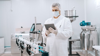 Person in weißem Kittel mit Haarnetz, Mundschutz und Gummihandschuhen hält Tablet in Händen und blickt darauf, vor ihr Halterung mit Dispensern, ringsum Laborgeräte