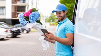 Lächelnde Person mit Kappe lehnt an Lieferwagen und blickt auf Tablet, in anderer Hand Blumenstrauß aus pinken und blauen Hortensien, im Hintergrund verschwommen Wohnhaus und parkende Autos