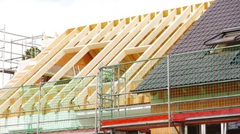 Auf einem Gebäude ist die eine Hälfte eines Dachs fertig. Die andere Hälfte des Dachs besteht aus schrägen Holzbalken. Innen sind Wände aus Ziegeln. Davor und links neben dem Gebäude steht jeweils ein Baugerüst