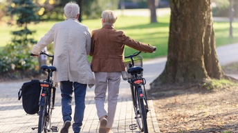 Rückenansicht zweier nebeneinander gehender Personen. Unter ihnen ist der Weg gepflastert. Beide Personen schieben neben sich ein Fahrrad. Im Hintergrund ist eine grüne Wiese mit Bäumen