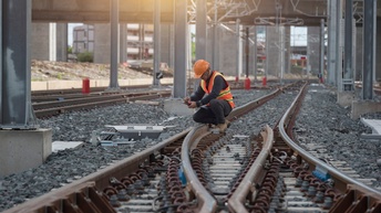 Eine Person arbeitet auf den Gleisen einer Bahnstrecke, sie hält ein Messgerät in den Händen. Im Hintergrund sieht man die Unterführung der Bahnstrecke. 