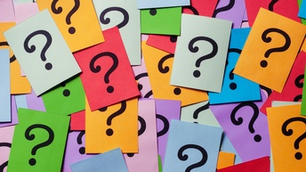 Topshot vieler kreuz und quer sowie teilweise übereinander liegender, bunter Zettel. Auf jedem Zettel ist ein schwarzes Fragezeichen