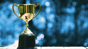 Goldener Pokal steht auf einem Holztisch, im Hintergrund zeigt sich ein blaues Bokeh, Konzept für Sieg