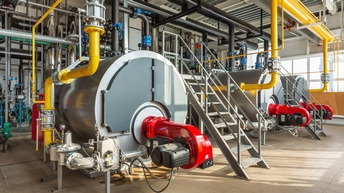 Drei große graue Kesselanlagen umgeben und verbunden mit zahlreichen gelben, schwarzen, roten und chromfarbenen Verrohrungen in Industriehalle