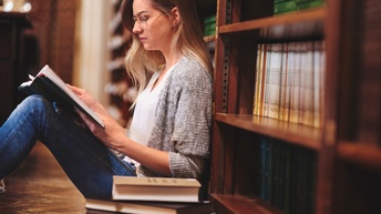 Person mit Brillen sitzt am Boden einer Bibliothek und liest in aufgeschlagenem Buch