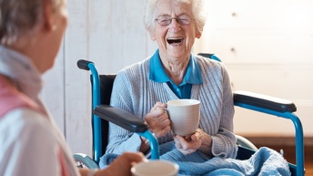 Lachende ältere Person mit Brillen sitzt in Rollstuhl und hält Tasse in der Hand, im Vordergrund verschwommen weitere Person mit Tasse