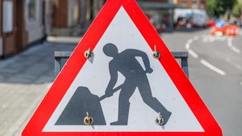 Auf einem dreieckigen Verkehrsschild ist die Silhouette einer Person, die eine Schaufel hält. Im Hintergrund ist eine mehrspurige Straße, auf der ein Teil mit orangen Vorrichtungen abgesperrt ist