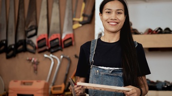 Porträt einer Person in einer Latzhose, die ein kleines Holzbrett und eine Säge hält. Im Hintergrund hängen an der Wand weitere verschiedene Sägen