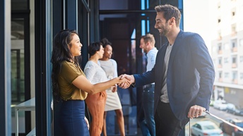Zwei Personen auf Balkon stehend reichen sich freundlich die Hände, im Hintergrund weitere Personen im Gespräch
