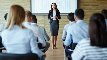 Person in Businesskleidung hält einen Vortrag, hinter ihr wird auf einer Leinwand eine Statistik projiziert und weitere Personen sitzen im Publikum