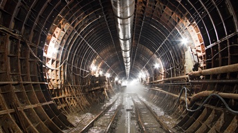 Ein breiter Gang wird rundum von abgerundeten Metallteilen umschlossen, sodass ein Tunnel entsteht. Am Boden sind vier Schienen, die nach hinten führen. Zwischen und unter den Schienen ist Schlamm