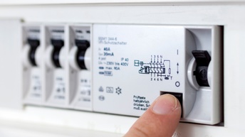 Nahaufnahme der Hand einer Person, die bei einem geöffneten Stromkasten den Fi-Schutzschalter drückt