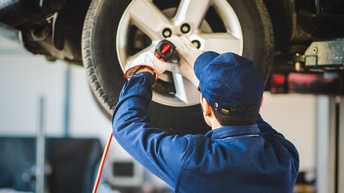 Rückenansicht einer Person in einem blauen Oberteil und einer blauen Kappe. Vor der Person schwebt ein Autoreifen. Die Person hält ein Gerät darauf. Das Gerät hat ein langes rotes Kabel. Die Person trägt Handschuhe und blickt darauf