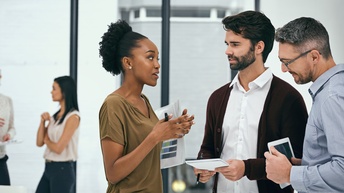 Drei Personen stehen in einem Halbkreis und interagieren miteinander. Eine Person hält ein Tablet, eine Person hält Unterlagen mit Statistiken und eine einen Notizblock. Im Hintergrund stehen zwei weitere Personen